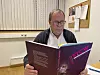 En mann med briller leser et lilla hefte ved et bord på et kontor.