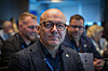 Mann med briller og mørk jakke foran en forsamling på et møte.