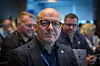 Mann med briller og mørk jakke foran en forsamling på et møte.