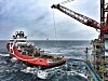 Rød og hvit supplybåt på urolig hav ved en offshoreinstallasjon med kran.