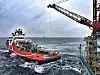 Rød og hvit supplybåt på urolig hav ved en offshoreinstallasjon med kran.