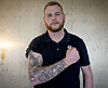 Skjeggete mann i mørk skjorte viser fram en tatovert underarm foran en lys vegg.