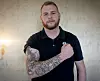 Skjeggete mann i mørk skjorte viser fram en tatovert underarm foran en lys vegg.