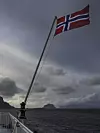 Norsk flagg vaier fra en ferge over mørkt hav, med fjell og skydekke i bakgrunnen.