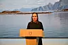 Kvinne taler bak en talerstol foran en fjord med fjell i bakgrunnen.
