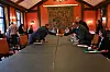 Personer sitter rundt et langt bord i et pyntet møterom under et formelt møte.